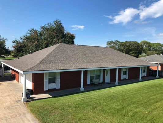 Red brick house with a gray roof and white columns, set on a green lawn under a blue sky. A driveway and carport are visible.