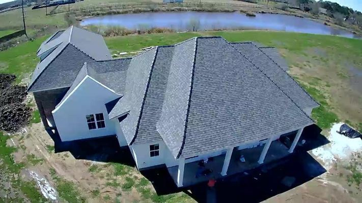 Aerial view of a white house with a dark gray roof, porch, and a pond in the background. The house is in a field.