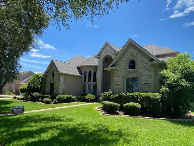 Large two-story house with light stone facade, gray roof, and lush green lawn under a blue sky.