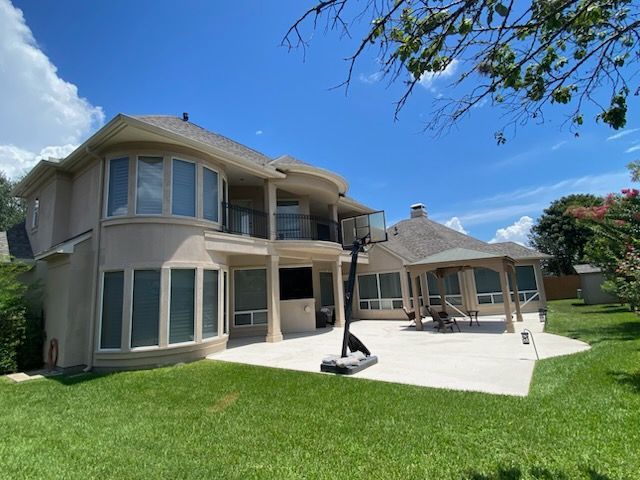 Two-story beige house with a patio, basketball hoop, and gazebo on a sunny day.