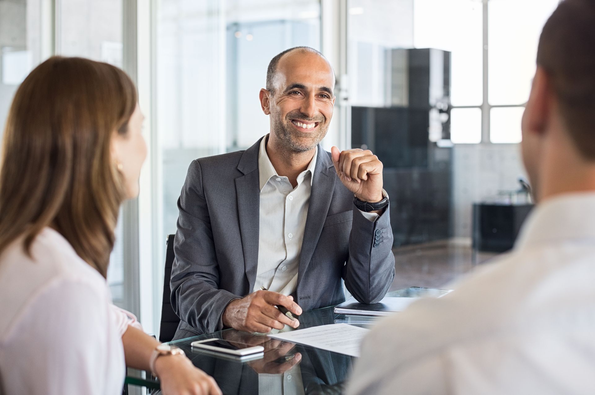 Business meeting with three people in a bright office, one man smiling and gesturing while seated at a table