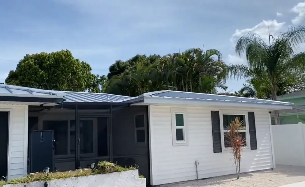 A white house with black shutters is surrounded by palm trees on a sunny day.