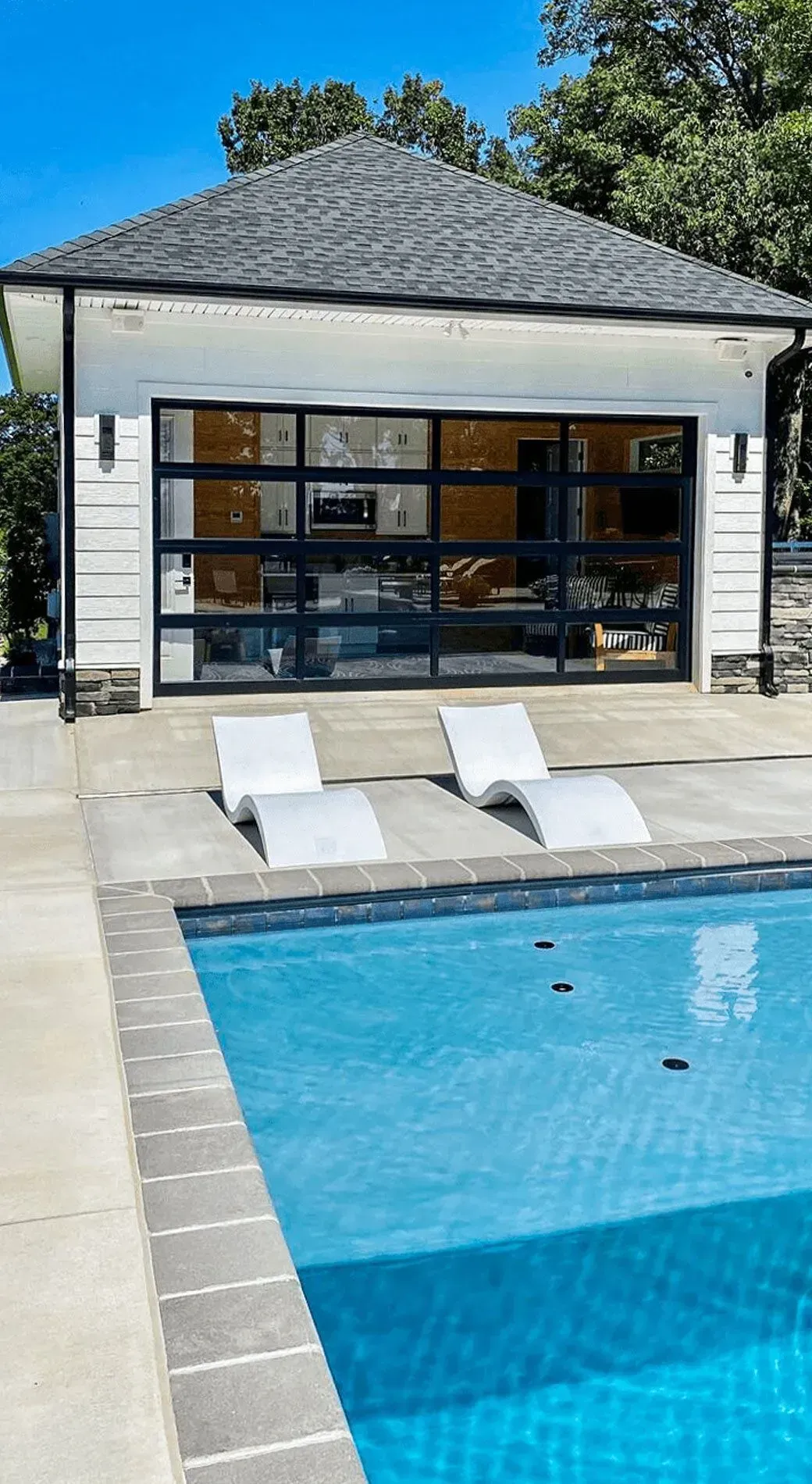 Swimming pool with white chaise lounges, in front of a building with black-framed glass doors.