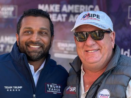 Two men are posing for a picture in front of a make America great again sign.