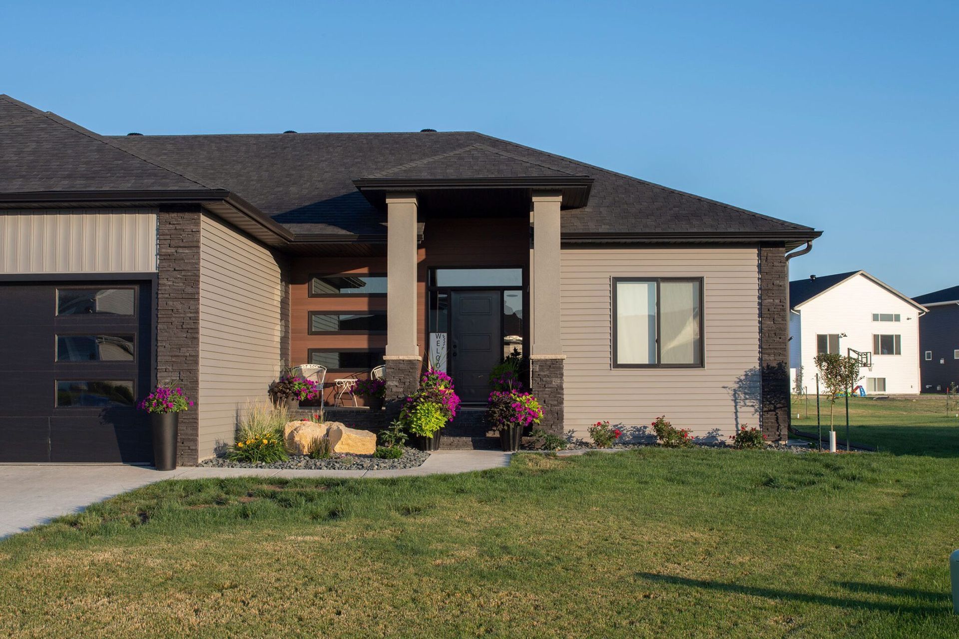 A modern, single-story suburban home with tan siding, dark brick accents, and a front porch, set in a sunny neighborhood.