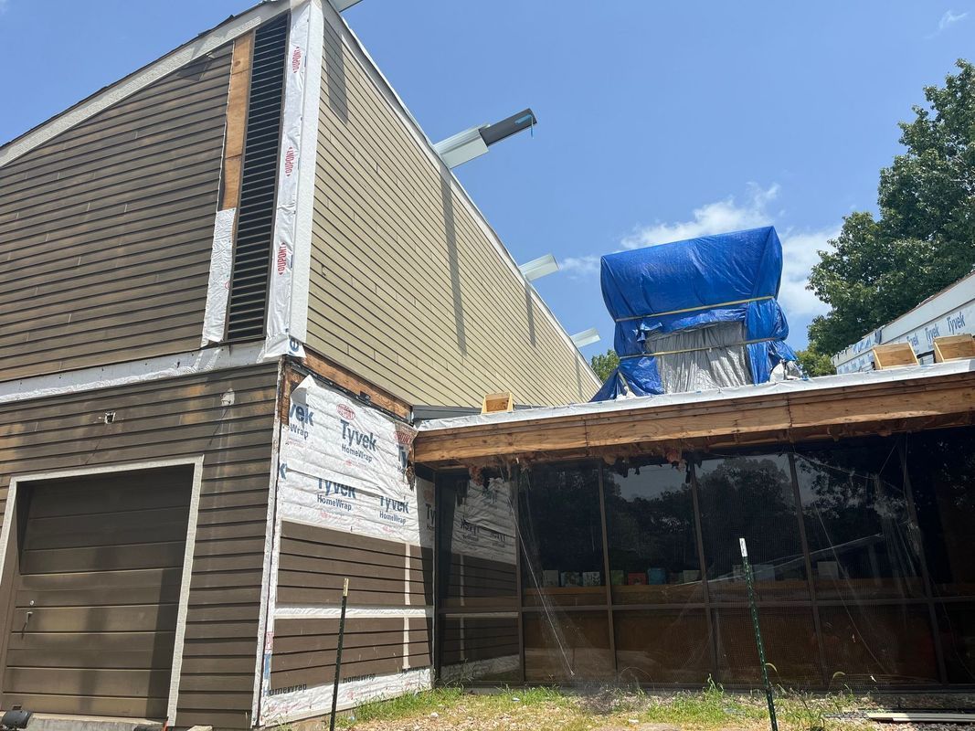 A house under renovation with partially removed siding, exposed house wrap, and a blue tarp covering part of the roof.