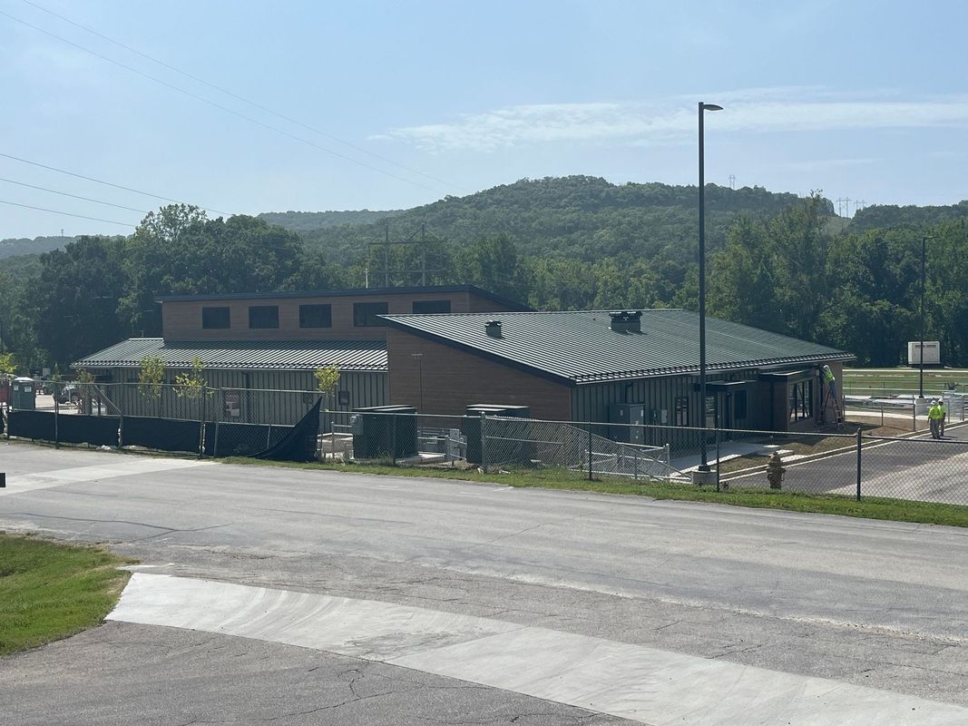 A low-slung brick building with a dark metal roof under construction, set against a backdrop of rolling, wooded hills.
