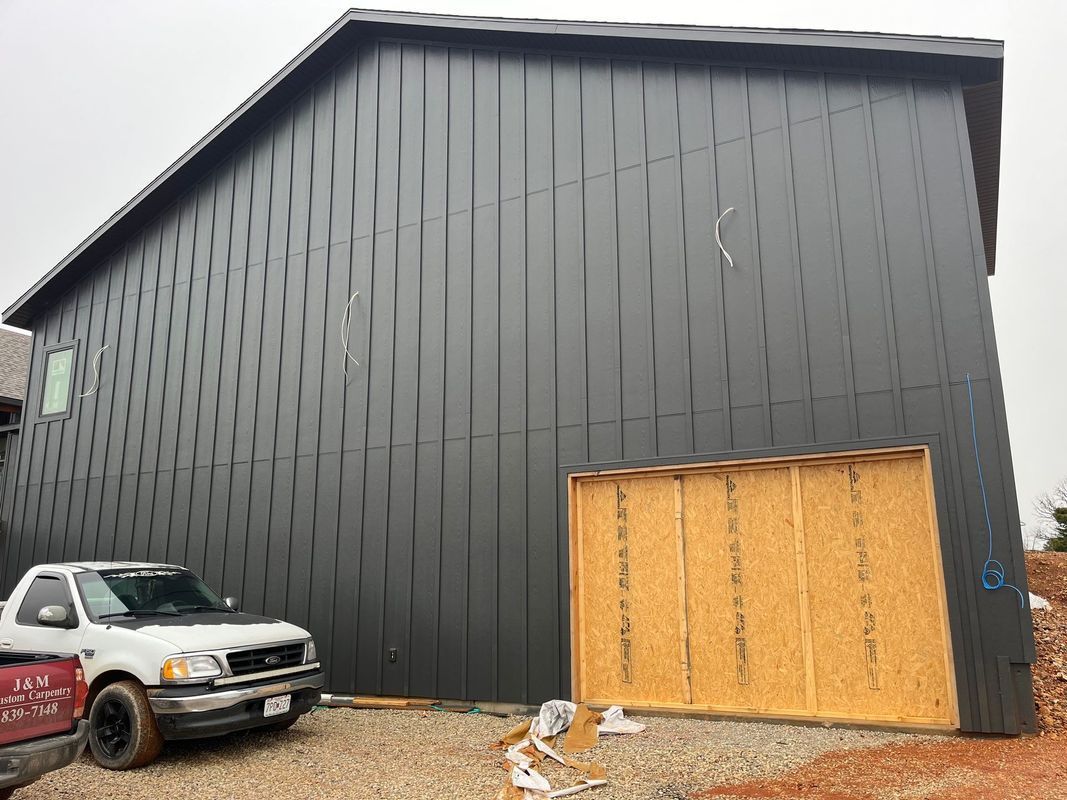 A construction site featuring a dark gray metal-sided building with a temporary wooden garage door cover and a parked truck.
