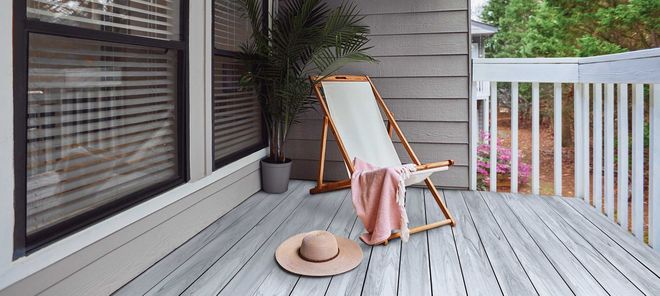 A lounge chair with a pink throw blanket and a sun hat sits on a gray wooden deck next to a potted palm and window.