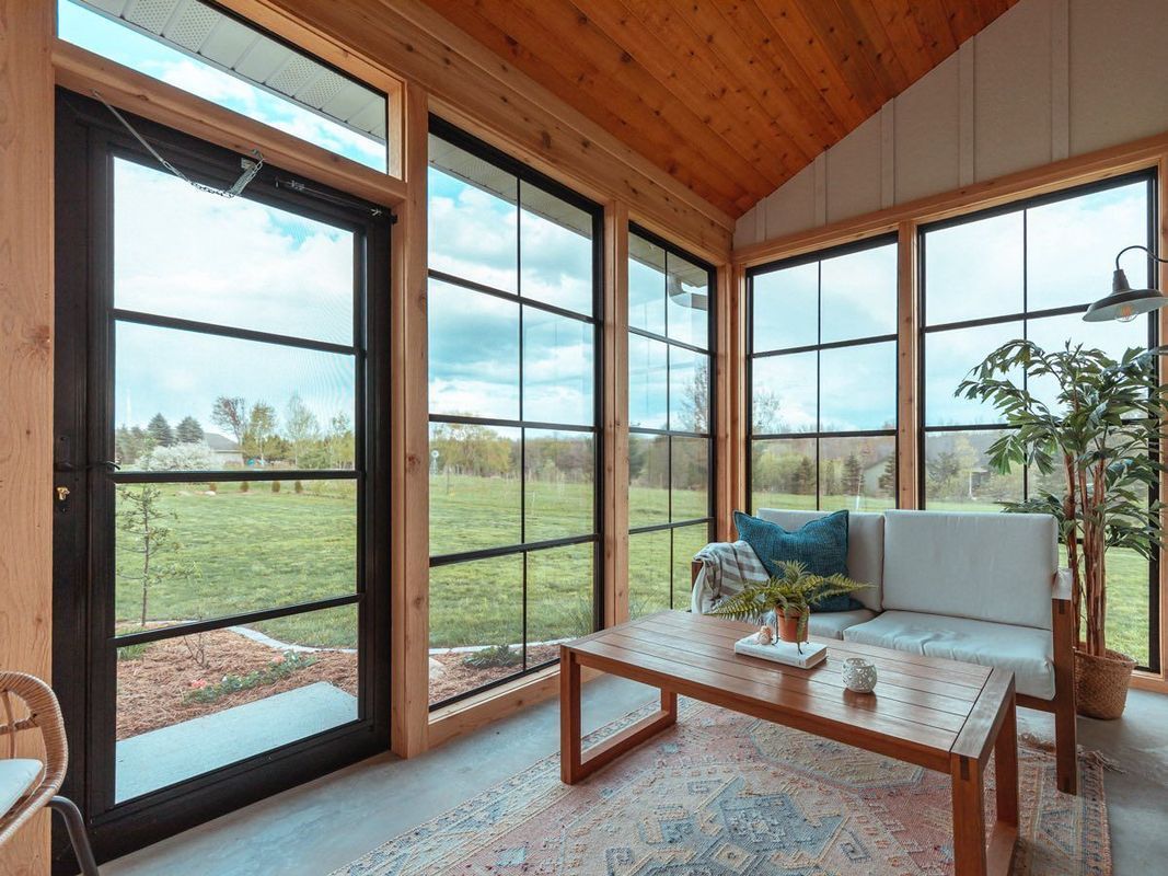 A sunroom with large windows and a glass door, featuring a wooden coffee table, a white sofa, and a potted plant.