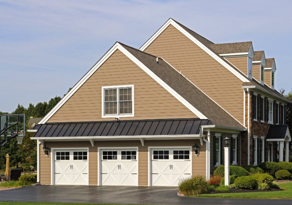 A tan suburban home with a three-car garage, a metal awning, and a paved driveway under a blue sky.