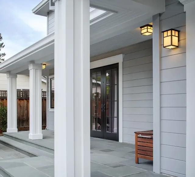 Gray, shingled porch entrance with columns, a stone floor, outdoor lanterns, and dark-framed French doors.