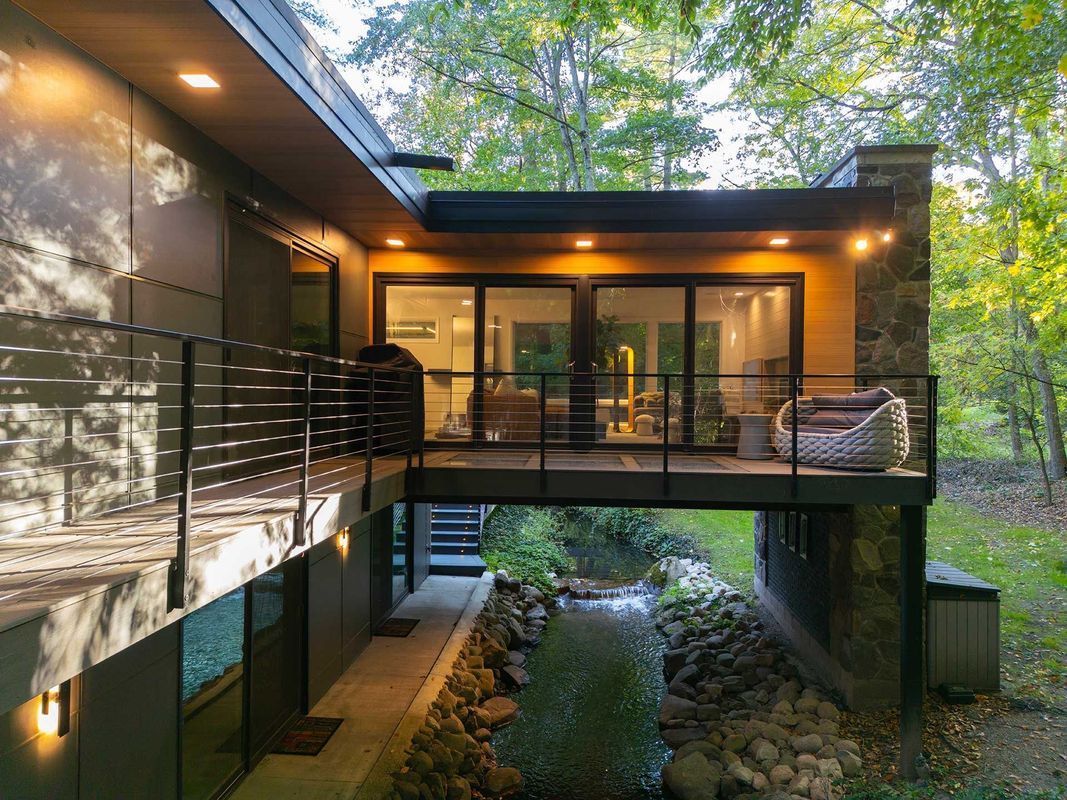 A modern home built over a creek, featuring a deck, glass walls, stone accents, and warm outdoor lighting.