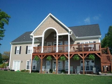 A two-story tan house with a large wooden deck, white porch columns, and a dark shingled roof under a clear blue sky.