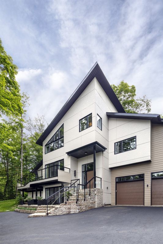 A modern three-story house with a white facade, a dark peaked roof, and a two-car garage, surrounded by lush green trees.