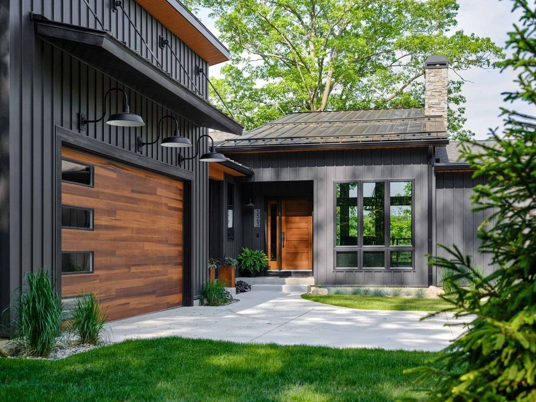 A modern home with dark vertical siding, a wood-paneled garage door, and a stone patio under a bright, sunny sky.
