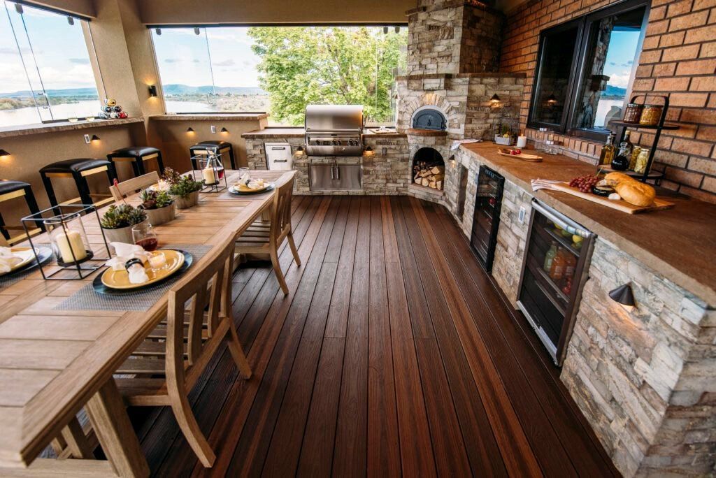A spacious outdoor patio featuring a wooden dining table, a stone grill station, a pizza oven, and a built-in refrigerator.