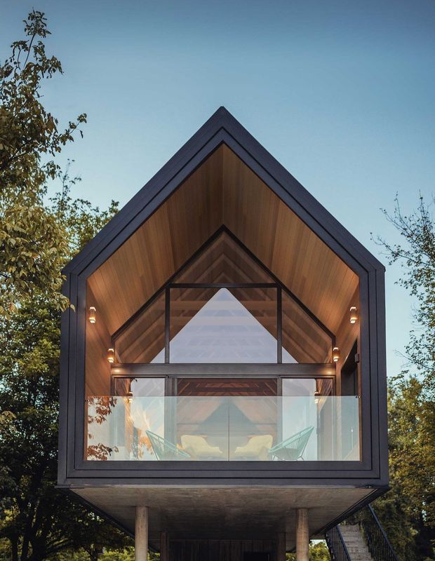 A modern A-frame house on stilts with a large glass balcony and wooden interior, surrounded by green trees.