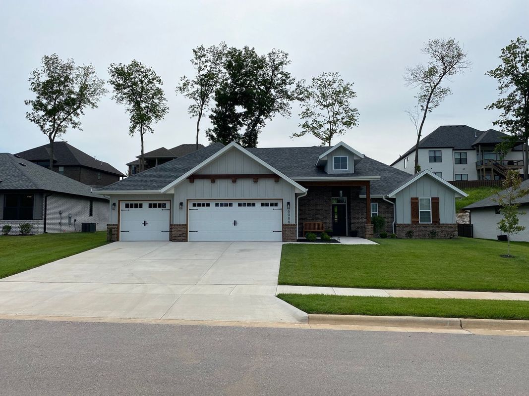 A single-story gray house with a stone facade, white garage doors, and a manicured lawn under a cloudy sky.