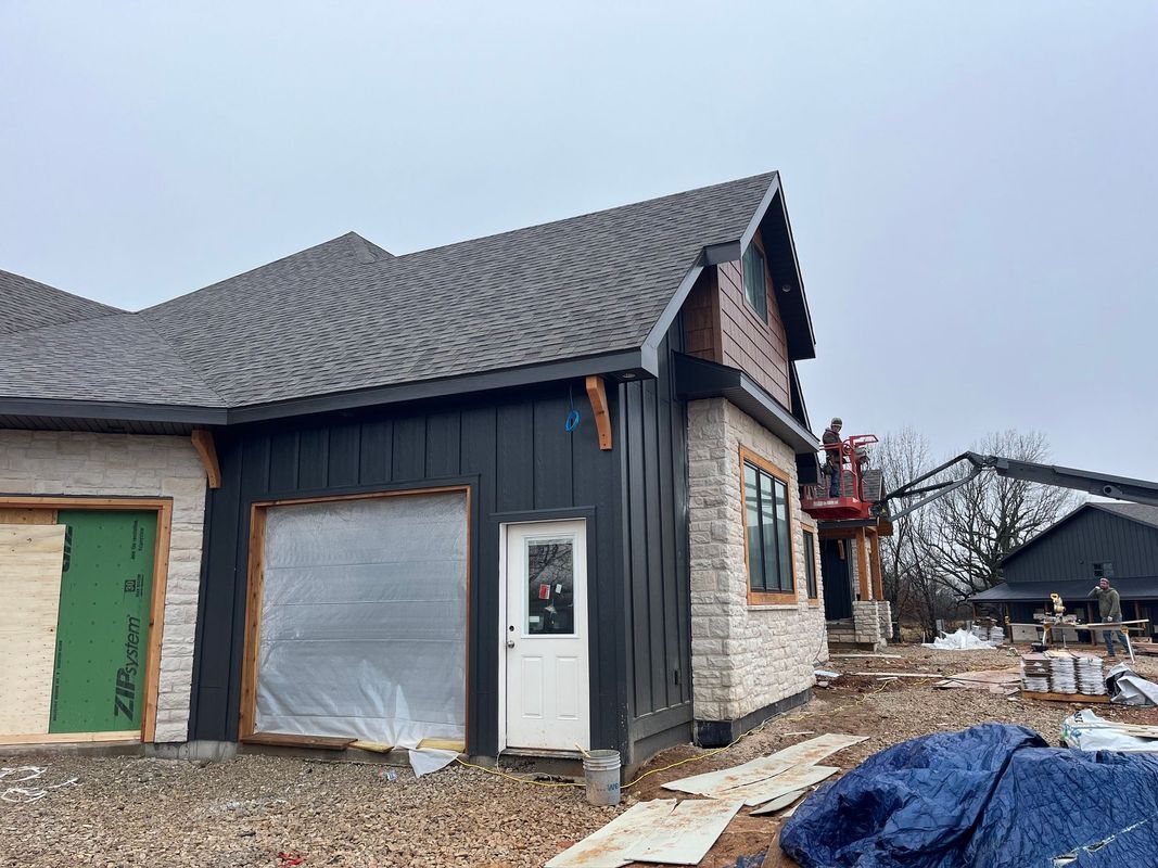 A partially constructed house exterior features black vertical siding, stone masonry, a shingled roof, and a white door.