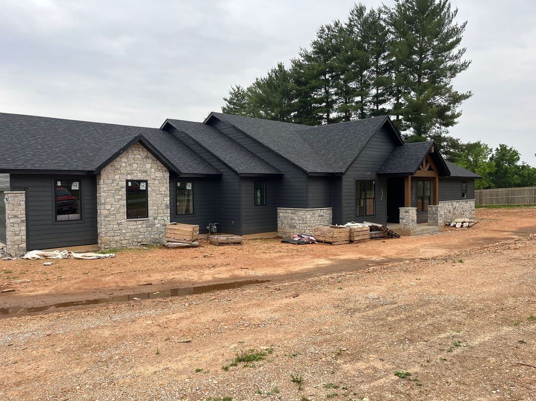 A single-story house under construction with dark siding, stone accents, and a black roof in a rural setting.