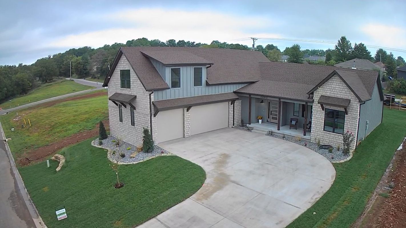An aerial view of a new two-story house with stone siding, a brown roof, and a large concrete driveway in a grassy yard.