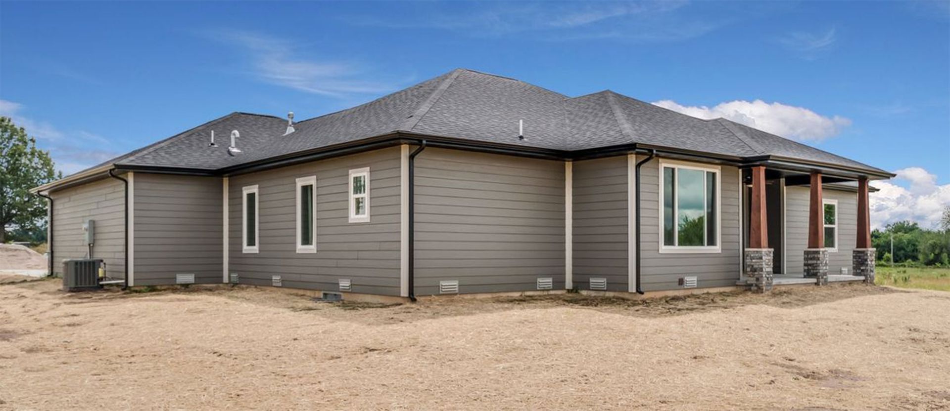 A modern gray single-story house with dark shingle roofing and wooden porch columns, set on a large, empty dirt lot.