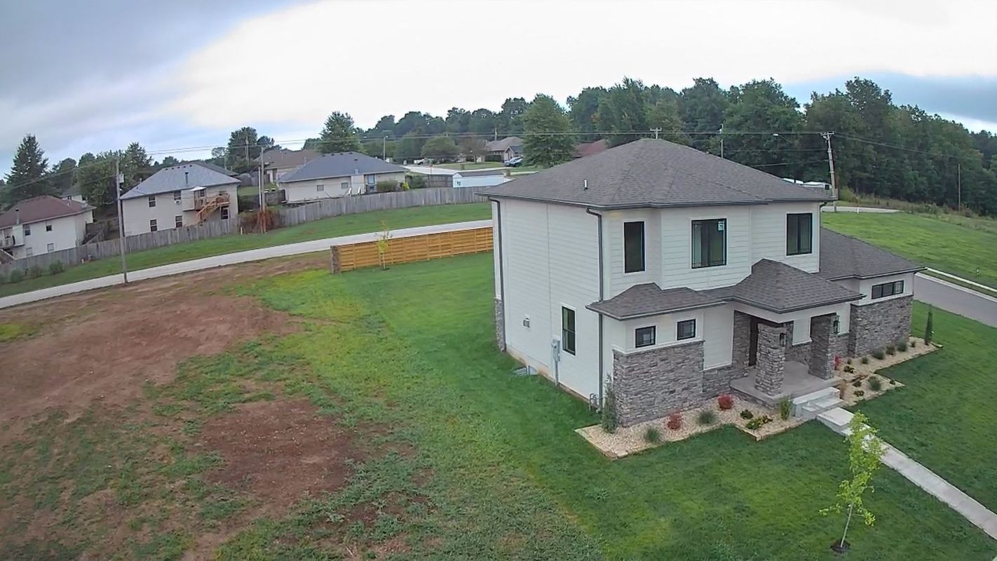 An aerial view of a two-story tan home with stone accents and a dark roof, situated on a grassy lot near a rural road.