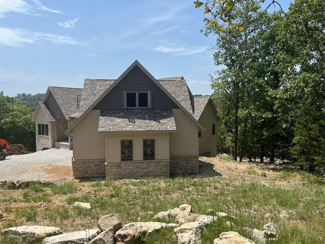 A tan and gray house with stone foundation set in a wooded, grassy landscape under a sunny, blue sky.
