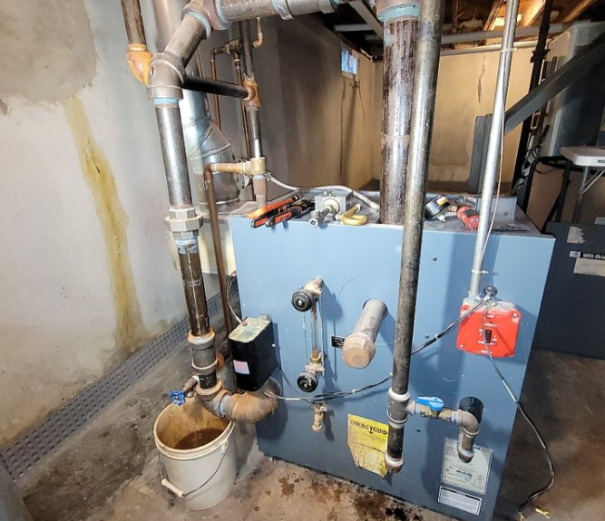 A blue furnace in a basement. Pipes, valves, and tools are present. A bucket collects brown water.