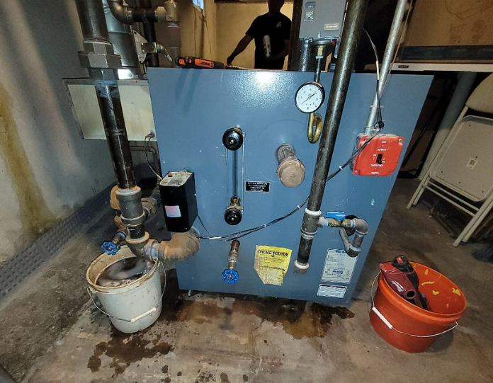 Blue boiler with pipes, gauges, and a person working in a basement. Buckets catch drips.
