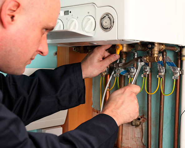 A man in a blue work suit is working on a boiler with a wrench.