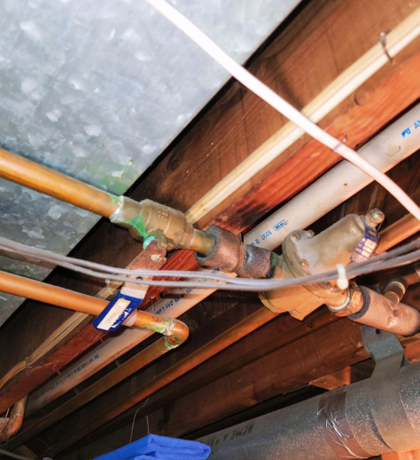 Copper and plastic pipes with valves under wooden beams, possibly in a basement.