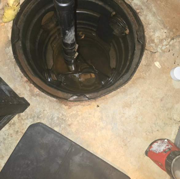 Overhead view of a dark sump pump basin with a pump and pipe. Concrete floor and surrounding items.