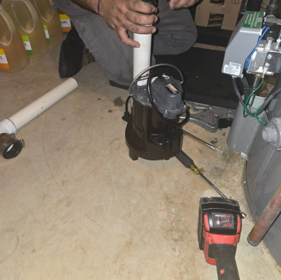 Person installing a sump pump, using a tool, near a furnace, in a basement setting.