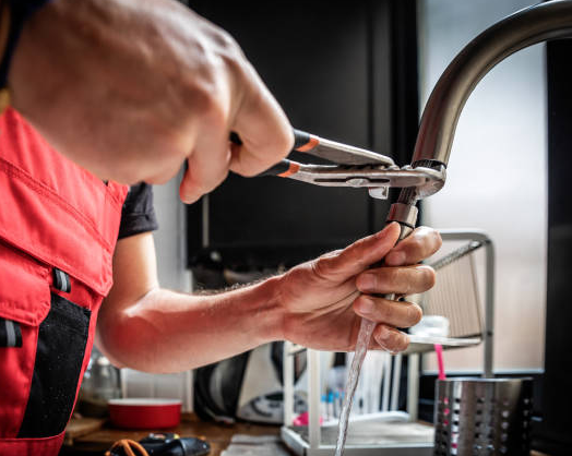 Plumber in red coveralls uses pliers on a faucet, water running.