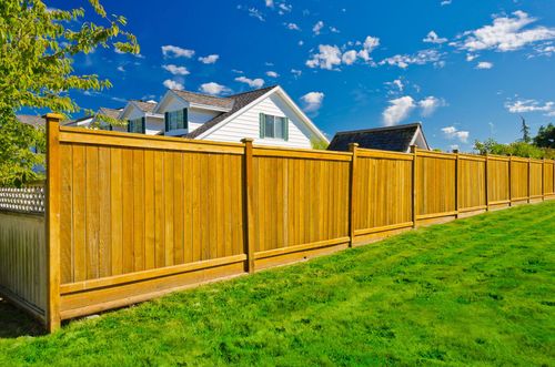 Wooden privacy fence beside a green lawn under a bright blue sky with a white house in the background