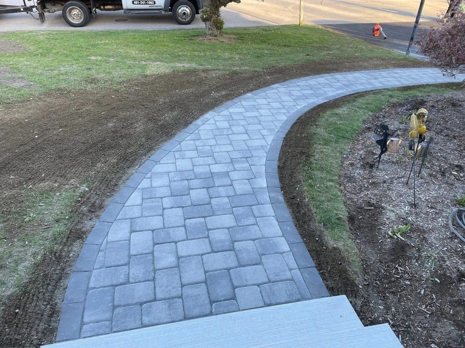 Curved gray paver walkway leading from a concrete step through a yard with mulch and grass.