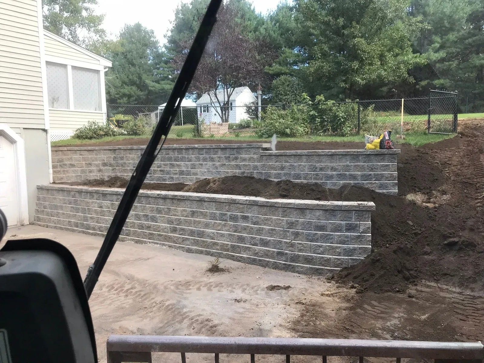 Backyard retaining wall under construction beside a house, with stacked stone blocks and exposed dirt slope.