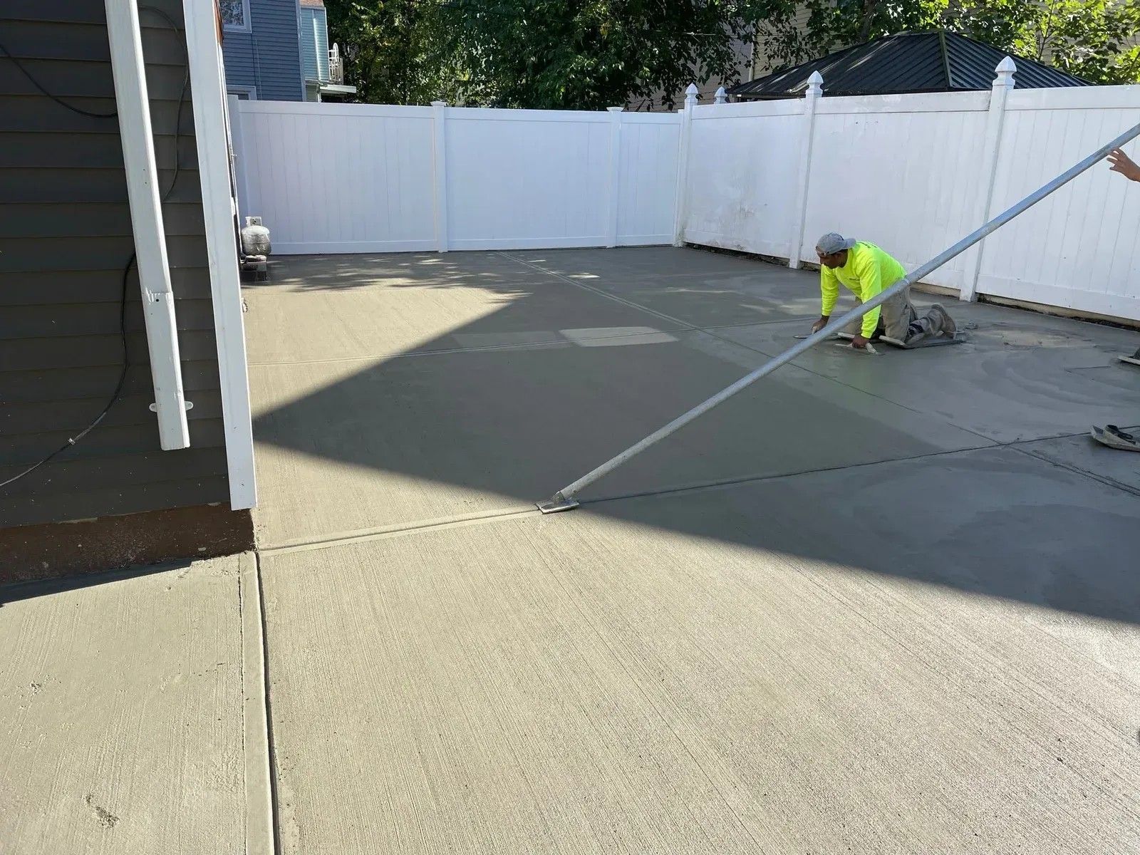 Worker in neon safety gear smoothing fresh concrete in a fenced backyard.