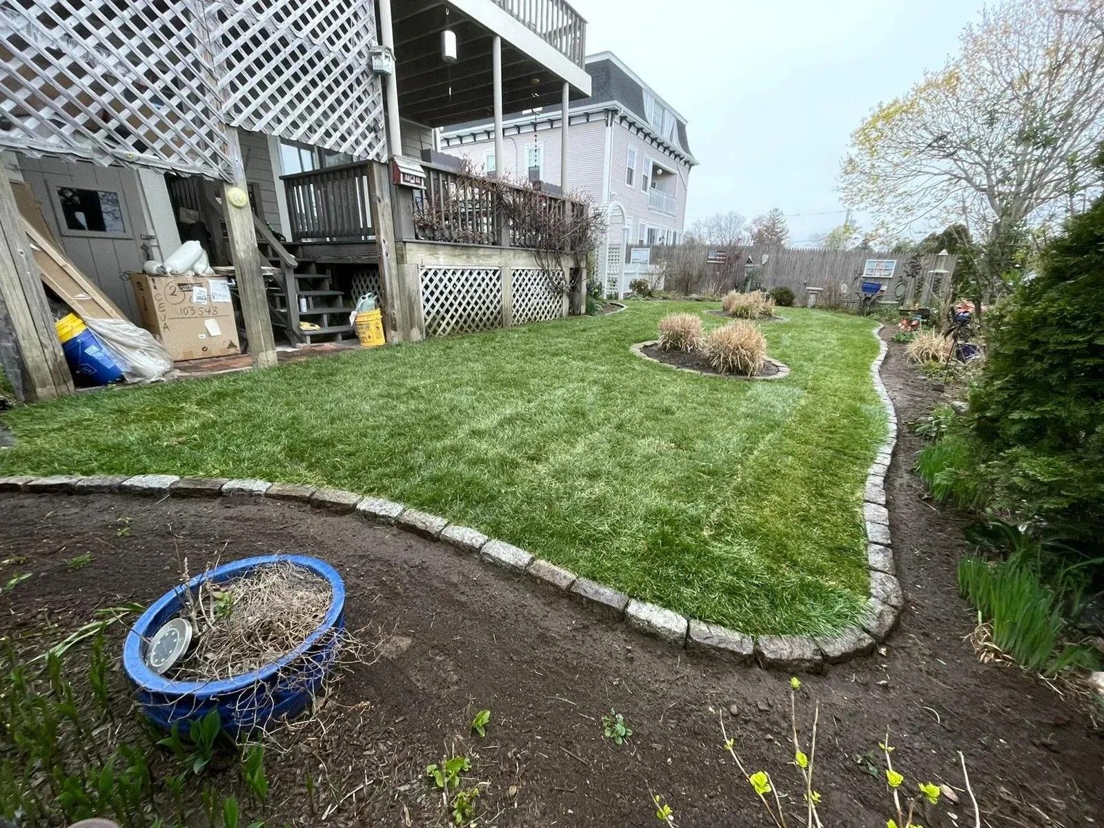 Backyard with green lawn, curved stone border, flower beds, and a house deck on the left.