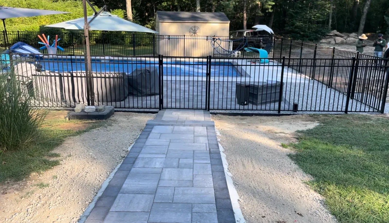 Stone path leading to a black fence and pool area with beige lounge chairs and umbrellas.