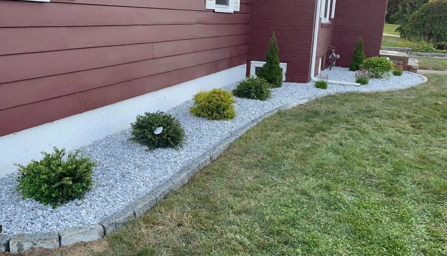 Landscaped side yard with small shrubs in white gravel beside a red house and green lawn.