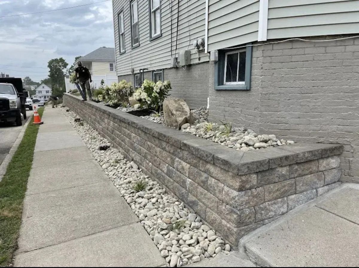 Stone retaining wall with white rock landscaping along a sidewalk beside a house.