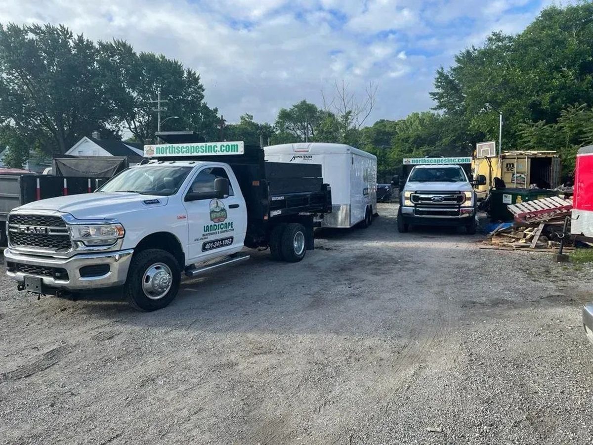 White work trucks and trailers parked on a gravel lot near trees and equipment under a cloudy sky.
