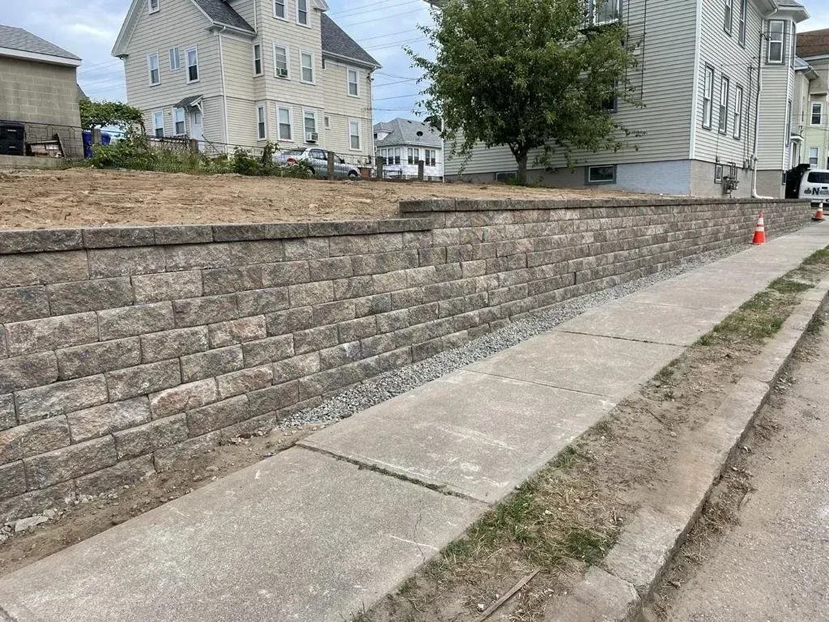 Stone retaining wall beside a sloped sidewalk in a residential neighborhood, with houses and an orange cone.