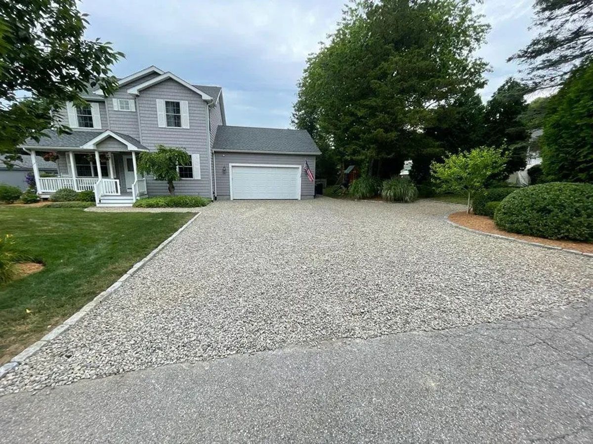 Gravel driveway leading to a gray two-story house with a garage and front porch, surrounded by green trees.
