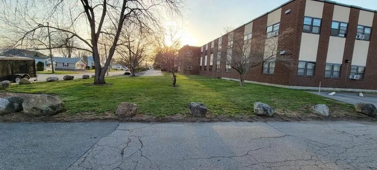 Brick building and grassy courtyard at sunset, with leafless trees and a cracked paved road in front.