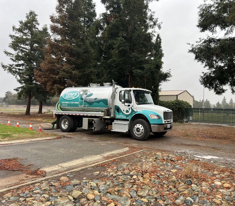 A tanker truck with teal and white paint parked on a cracked road in front of houses under a blue sky.