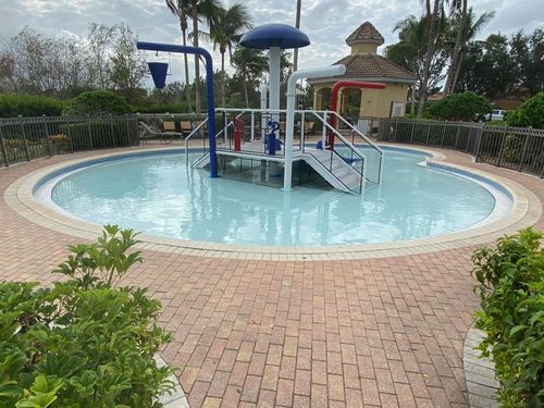 Children&rsquo;s splash pool with blue water, play structures, and brick deck in a landscaped park